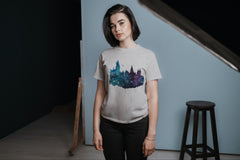 A young woman standing in front of a stool wearing a Hogwarts Castle Tshirt in greys, greens, blues, and purples.