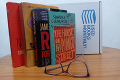 A stack of used books in front of a Used Books Monthly subscription box showcasing a book subscription service
