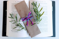 A bookmark wrapped in brown craft paper and decorated with ribbons rests on an open hardcover book. The bookmark is handmade and painted.