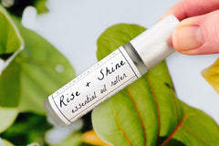 Two fingers holding the Rise + Shine Essential Oil roller bottle with an array of green foliage in the background.