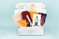Open box of IndieStitch with contents sitting in the box: such as, rolls of fabric, postcard, sewing notions, and a photo of the Roxy Jumper