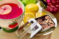 The Sipping Societea Loose leaf tea subscription tea tin with raspberry lemonade herbal tea next to decorative tea cup and lemons.