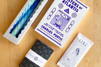 December 2023 box: Masters of Atlantis by Charles Portis. Contains "Cult" sea salt chocolate, spiral candles, and letterpress notebook.