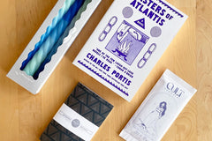 December 2023 box: Masters of Atlantis by Charles Portis. Contains "Cult" sea salt chocolate, spiral candles, and letterpress notebook.