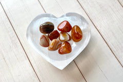 A photo of a heart-shaped selenite bowl holding several carnelians tumbled stones as an example of a crystal of the month.