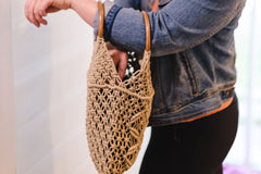 A woman in a denim jacket reaches into a woven macrame tote bag with round wooden handles, hanging from her arm.