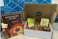 A Vietnamese coffee phin, a package of chai, a winter drinks of the world recipe booklet, a Chinese wine cup, and Stranger's Guide: Vietnam