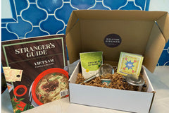 A Vietnamese coffee phin, a package of chai, a winter drinks of the world recipe booklet, a Chinese wine cup, and Stranger's Guide: Vietnam