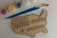 United States wooden shape with the words "United States of America" with a flag pattern. There are two paint brushes and a paint sampler.