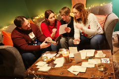 Group of friends play the story together with fairylights behind.  table shows a game in progress with a variety of letters and postcards
