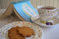 the image shows an open book, reading glasses, cup of tea and a plate containing 3 biscuits
