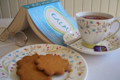 the image shows an open book, reading glasses, cup of tea and a plate containing 3 biscuits