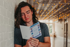 young adult male leaning up against a white brick wall while reading a can't stop won't stop thinking of you greeting card