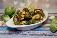 A white dish holding lemon garlic roasted brussels sprouts, garnished with sliced roasted garlic. Raw brussels & garlic are next to it.