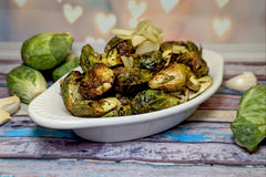 A white dish holding lemon garlic roasted brussels sprouts, garnished with sliced roasted garlic. Raw brussels & garlic are next to it.