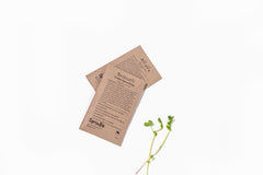 Two seed packets sit on top of each other with a white background. They are brown paper. One says "alfalfa" and the other "broccoli".