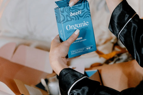 A unboxing image with a woman holding a box of organic tampons. In the background are other items from the box like self care products.