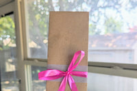 A Romance-themed bookmark is wrapped in brown craft paper and decorated with pink ribbon. It rests on an open hardback book.