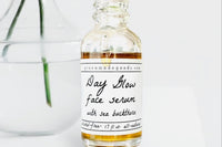 The Day Glow Serum by Greenmade goods. A bottle with golden serum dripping down the label. A clear vase with water in the background.