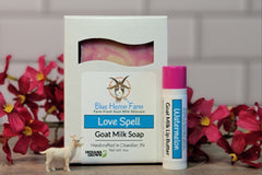 Love Spell special February scent goat milk soap bar with a Watermelon Lip Butter sitting on a table surrounded by pink flowers.