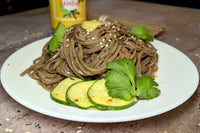 A white plate with a delicious-looking pile of soba noodles. It is garnished with marinated cucumber, cilantro leaves, sesame seeds & sambal