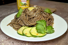 A white plate with a delicious-looking pile of soba noodles. It is garnished with marinated cucumber, cilantro leaves, sesame seeds & sambal