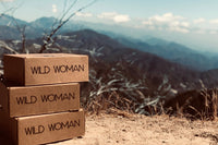 Wild Woman Box subscription boxes stacked on a mountain ledge with a scenic view in the background
