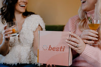 Two women pictured together drinking cocktails and smiling. They are holding a Besti box between them. 