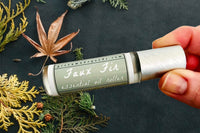 Two fingers holding the Faux Fir Essential Oil roller bottle with dried leaves and fall foliage in the background.