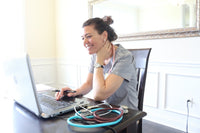 Nurse with dark hair and scrubs on sitting at computer in office with stethoscopes on table ordering on the computer and smiling