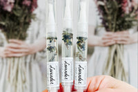 Hand Holding Three Lavender Botanical Nail Pens by Greenmade goods with Ladies in White Dresses Holding Bouquets in the Background.