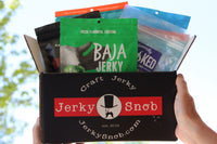Black logo box with 8 bags of jerky.  Baja Lime Serano, Honestly Smoked Mild Child, Savage Teriyaki, Baja Peppered, Werner BBQ.
