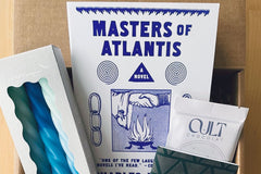 December 2023 box: Masters of Atlantis by Charles Portis. Contains "Cult" sea salt chocolate, spiral candles, and letterpress notebook.