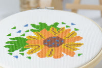 counted cross stitch beginner friendly craft of orange and yellow flower with green leaves on white embroidery fabric inside wood hoop