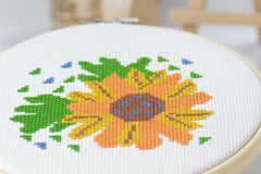 counted cross stitch beginner friendly craft of orange and yellow flower with green leaves on white embroidery fabric inside wood hoop