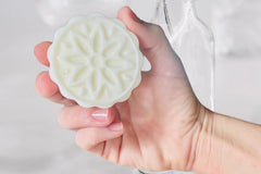 Hand holding the Sandalwood Lotion Bar by Greenmade goods, showing it's molded flower pattern with wood slats and plants in the background.