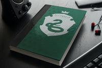 Green hard cover Slytherin Journal sitting on a desktop next to a computer speaker, keyboard, and earbuds.