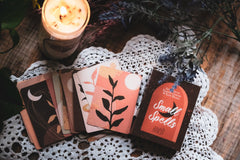 deck of cards fanned out next to a lit candle on a white doily next to sprigs of purple flowers all on a wood table