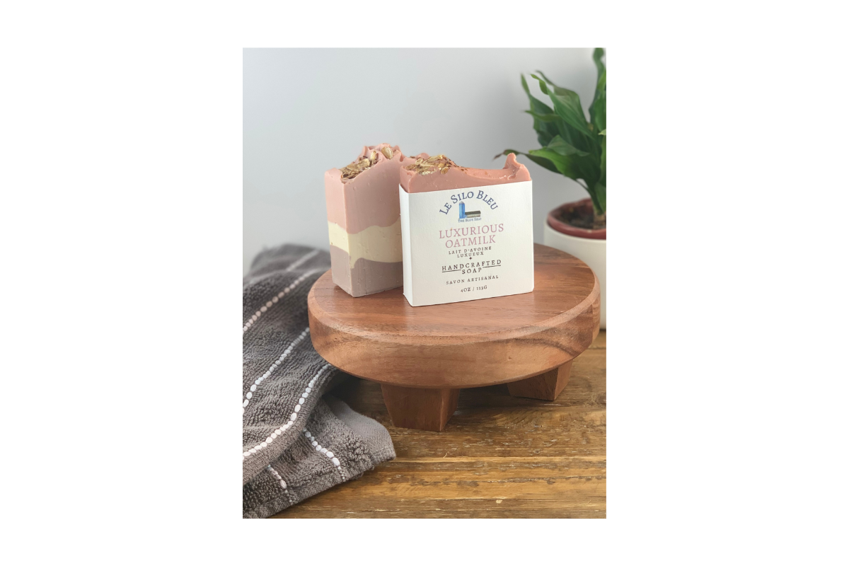 Pink, purple and while layered bars of soap on top of a wooden stand with a packaged soap and a towel and plant in the background.