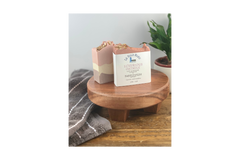 Pink, purple and while layered bars of soap on top of a wooden stand with a packaged soap and a towel and plant in the background.