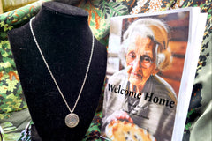 Short story titled Welcome Home with old woman holding a pie on the cover. There is also a silver necklace with a crescent moon and leaves.