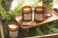 two jarred candles sit on a wooden riser with a box of matches and a plant. another candle sits on the table adjacent. 