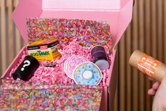 Someone holds a pink box containing 4 rolls of film, as well as two stickers of a donut and a pin, packed with pink crinkle paper