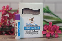 Picture of a past Soap Of The Month, "Lilacs In Bloom". Purple soap in a white box next to a "Very Berry" Lip Butter and pink flowers.