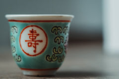 A ceramic Chinese wine cup. It is small, light blue with red highlights and Chinese characters and other decorative features. 