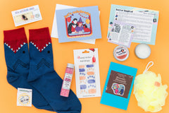 A scary movie-themed subscription box. Items include shark socks, bath salts, dark humor greeting card, enamel pin, notebook, and candle.