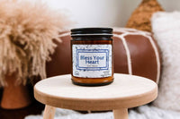 a jarred candle named bless your heart sits on a small wooden stool. a tufted leather footstool is in the background, as well as some pillow