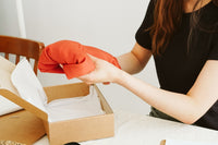 Woman in a black dress and brown hair is sitting at a table. She is removing a piece of clothing from a box. 
