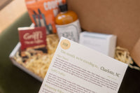 A hand holding a white cap with green text saying "shop small, live well, explore local" for the Explore Local Box monthly subscription