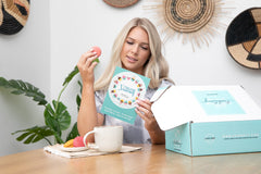 blond woman reading the insert card of a Bath Bevy Subscription box while eating a macaroon in a boho setting